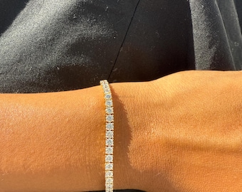 May include: A gold tennis bracelet with a line of round, clear gemstones. The bracelet is worn on a person's wrist, with a black shirt in the background. The gemstones are set closely together, creating a continuous sparkle.