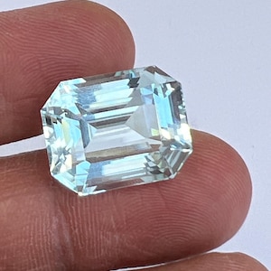 May include: A rectangular, aquamarine gemstone with an emerald cut. The gem is held between fingers, showcasing its light blue and clear facets. The stone's cut reflects light, creating a sparkling effect.