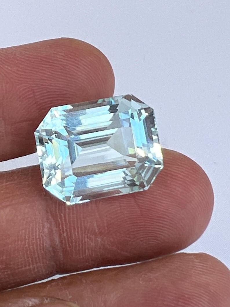 May include: A rectangular, aquamarine gemstone with an emerald cut. The gem is held between fingers, showcasing its light blue and clear facets. The stone's cut reflects light, creating a sparkling effect.
