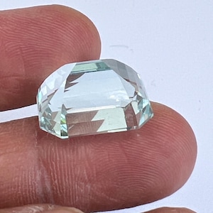 May include: A faceted, light blue gemstone with a rectangular shape and a domed top. The gem is held between two fingers, showcasing its clarity and multiple facets that reflect light. The gem's color is a pale, translucent blue.