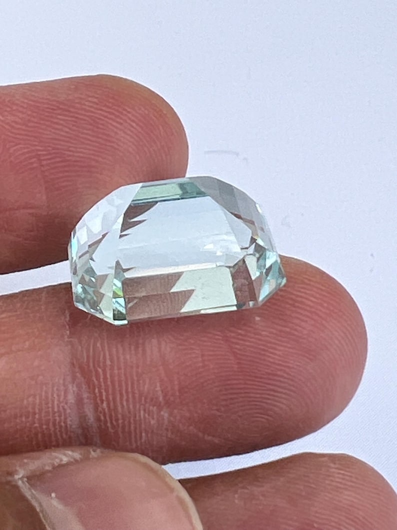 May include: A faceted, light blue gemstone with a rectangular shape and a domed top. The gem is held between two fingers, showcasing its clarity and multiple facets that reflect light. The gem's color is a pale, translucent blue.