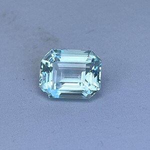 May include: A rectangular, light blue gemstone with an emerald cut. The gem has multiple facets that reflect light, creating a sparkling effect. The stone is set against a light gray background.