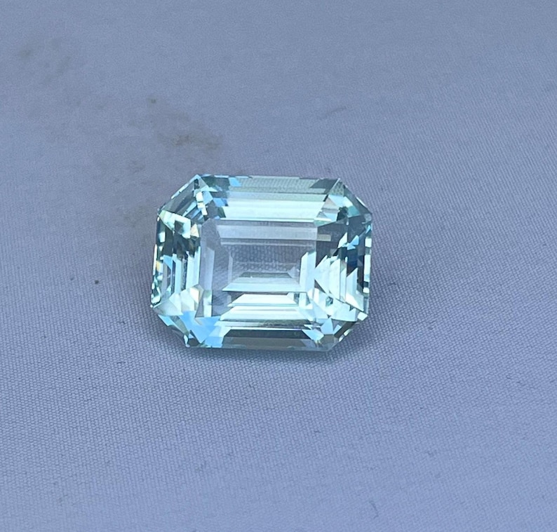 May include: A rectangular, light blue gemstone with an emerald cut. The gem has multiple facets that reflect light, creating a sparkling effect. The stone is set against a light gray background.