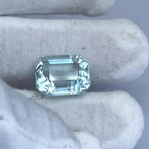 May include: A rectangular, light blue gemstone with multiple facets. The gemstone is held against a white fabric background. The gem has a clear, transparent appearance and reflects light.