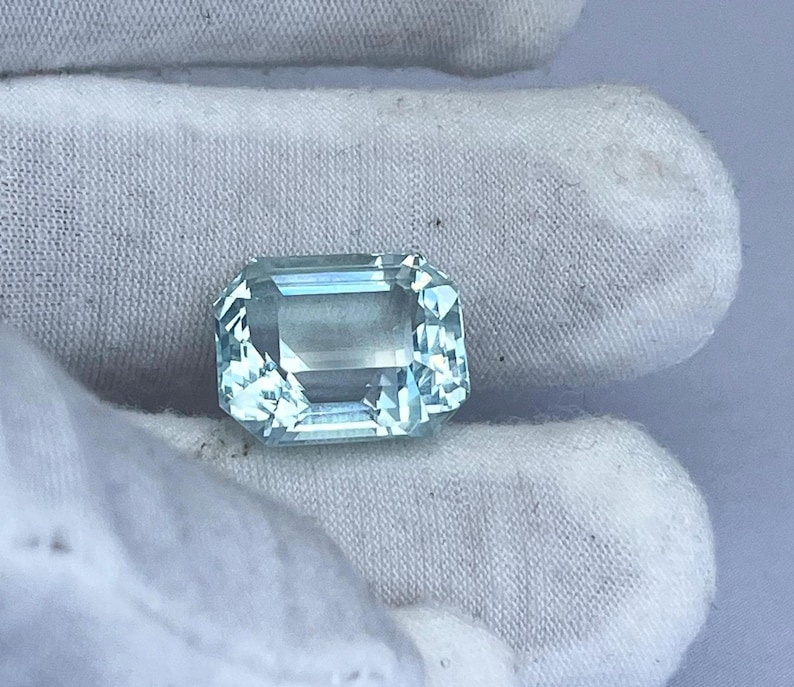May include: A rectangular, light blue gemstone with multiple facets. The gemstone is held against a white fabric background. The gem has a clear, transparent appearance and reflects light.