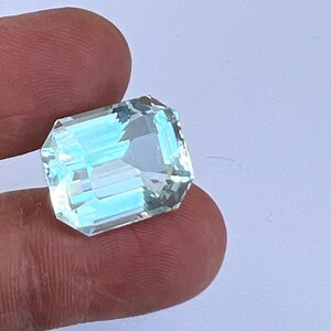 May include: A rectangular, faceted aquamarine gemstone with a light blue hue. The gem is held between fingers, showcasing its clarity and cut. The gemstone's facets reflect light, creating a sparkling effect. This gemstone is ideal for jewelry making.