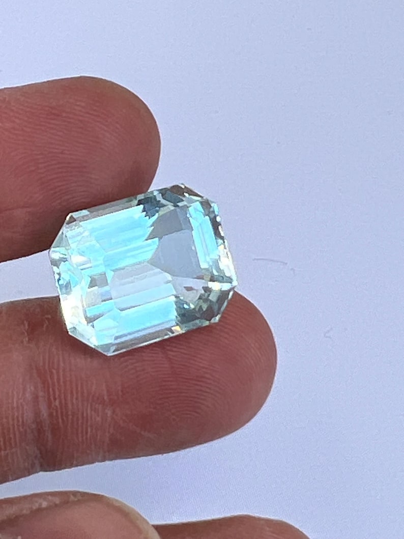 May include: A rectangular, faceted aquamarine gemstone with a light blue hue. The gem is held between fingers, showcasing its clarity and cut. The gemstone's facets reflect light, creating a sparkling effect. This gemstone is ideal for jewelry making.