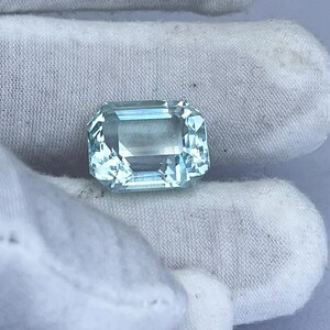 May include: A rectangular, light blue gemstone with multiple facets. The gemstone is held by a white fabric. The gem has a clear, transparent appearance, reflecting light. This gemstone could be used in jewelry.