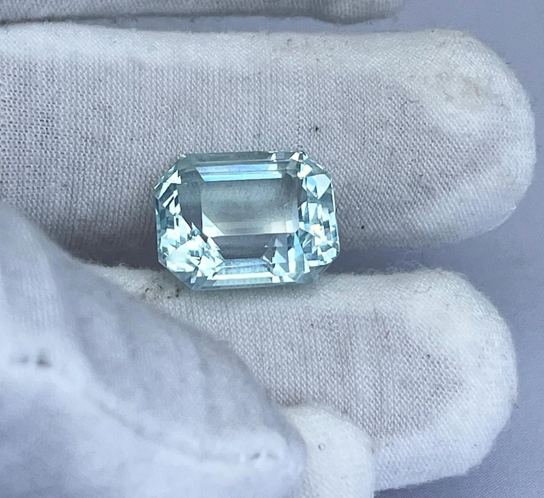 May include: A rectangular, light blue gemstone with multiple facets. The gemstone is held by a white fabric. The gem has a clear, transparent appearance, reflecting light. This gemstone could be used in jewelry.
