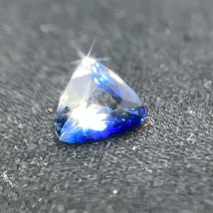 Op de afbeelding: Een driehoekige edelsteen met een briljante slijpvorm, met een kleurengradatie van helder tot diepblauw. De edelsteen is geplaatst tegen een donkere, getextureerde achtergrond, die de facetten en de glans benadrukt. De vorm van de edelsteen doet denken aan een driehoek.