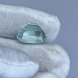 May include: A faceted, light blue gemstone with a geometric cut. The gem is held against a white fabric background. The gemstone's facets reflect light, creating a sparkling effect. The overall shape is a modified octagon, showcasing the gem's clarity and color.