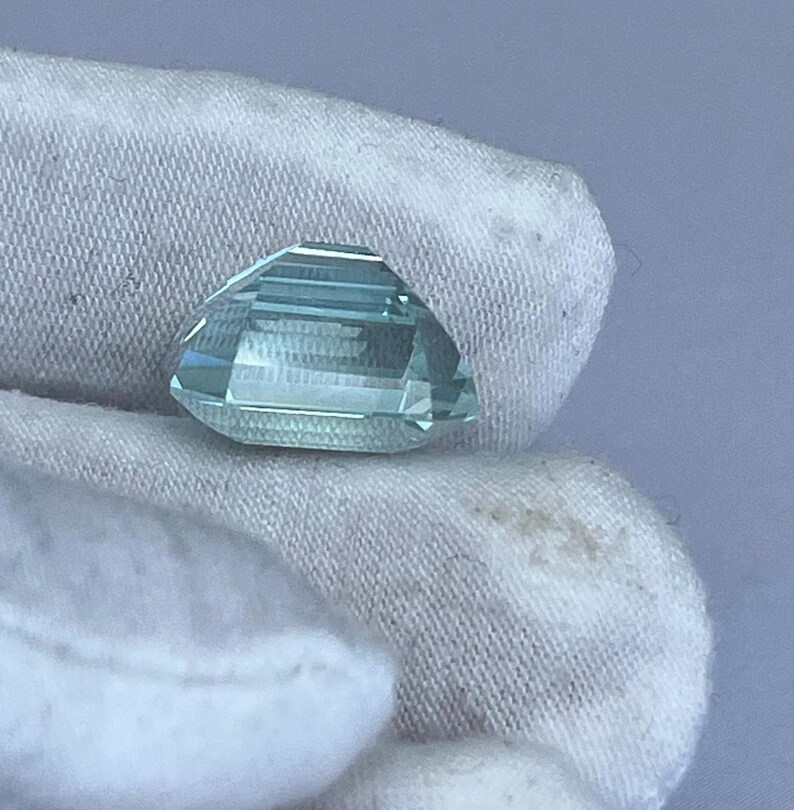 May include: A faceted, light blue gemstone with a geometric cut. The gem is held against a white fabric background. The gemstone's facets reflect light, creating a sparkling effect. The overall shape is a modified octagon, showcasing the gem's clarity and color.
