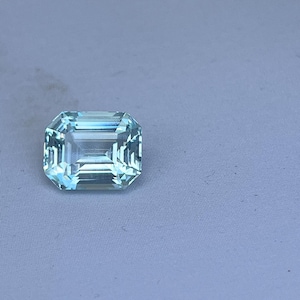 May include: A rectangular, light blue gemstone with a faceted surface. The gem has a clear, transparent appearance and is set against a light blue background. The cut of the gem is an emerald cut, with a series of parallel facets.