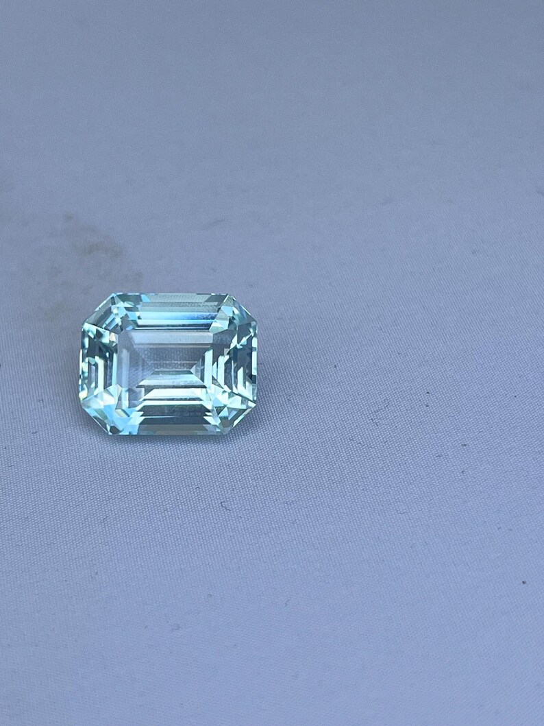 May include: A rectangular, light blue gemstone with a faceted surface. The gem has a clear, transparent appearance and is set against a light blue background. The cut of the gem is an emerald cut, with a series of parallel facets.