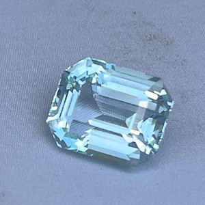May include: A faceted, light blue gemstone with an emerald cut. The gem is rectangular with cut corners and a transparent appearance. The stone is set against a light gray background.