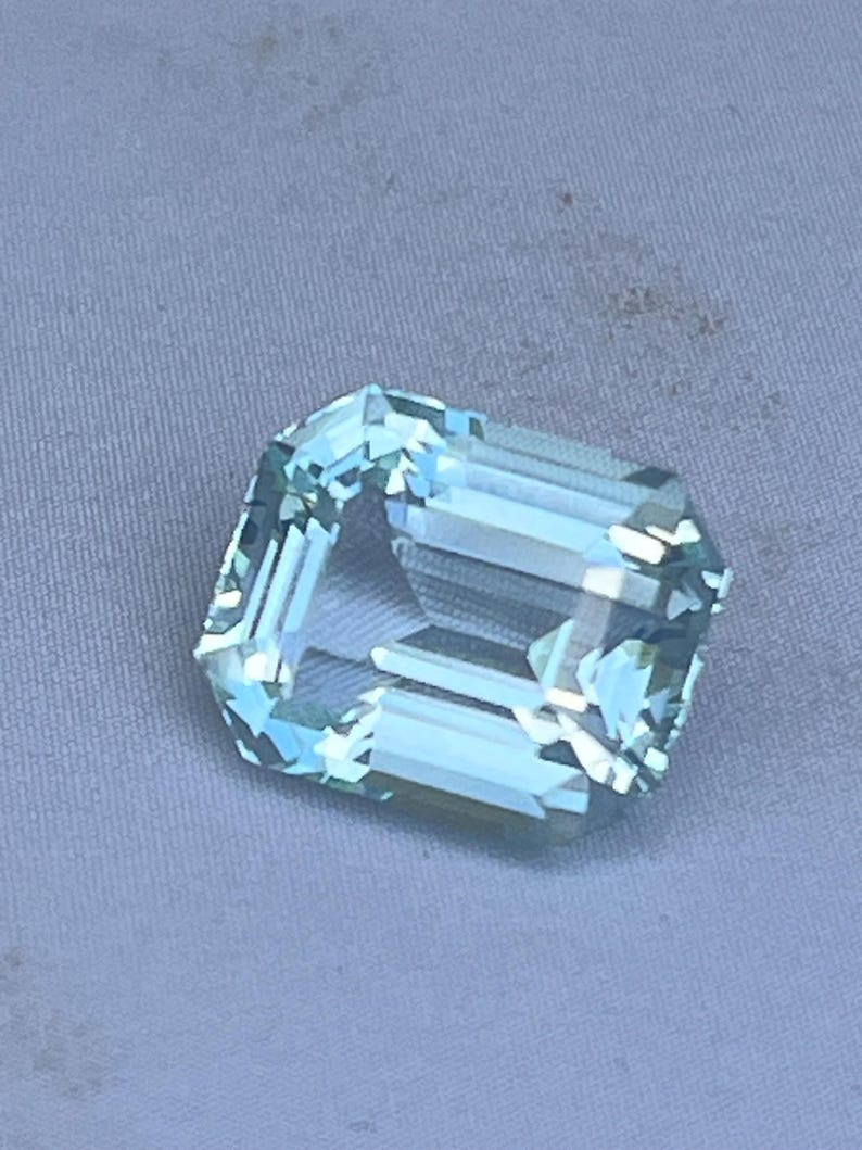 May include: A faceted, light blue gemstone with an emerald cut. The gem is rectangular with cut corners and a transparent appearance. The stone is set against a light gray background.