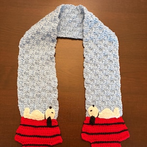 May include: A light blue crocheted scarf featuring a repeating pattern. The scarf has red and black Snoopy designs at each end. The Snoopy designs are wearing red shirts with black stripes and have white bodies and black ears.