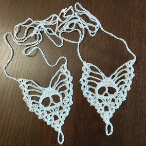 May include: White crochet barefoot sandals with a skull design. The sandals feature a toe loop and intricate openwork patterns. The delicate, handmade design is displayed on a dark brown surface.