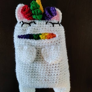 May include: White crocheted unicorn stuffed animal with a rainbow mane and horn. The unicorn has a small mouth and closed eyes.