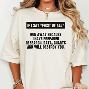 May include: Cream-colored t-shirt with black text. The text reads: "IF I SAY 'FIRST OF ALL' RUN AWAY BECAUSE I HAVE PREPARED RESEARCH, DATA, CHARTS AND WILL DESTROY YOU."