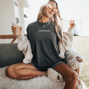 May include: A woman wearing a dark grey t-shirt with the text "cool mom" printed on it. She is sitting on a couch with a white throw blanket and holding a glass of coffee.