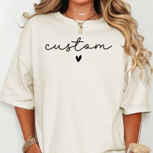 May include: Cream-colored t-shirt with the word "custom" in a black script font and a small black heart below. The shirt has a relaxed fit and short sleeves.
