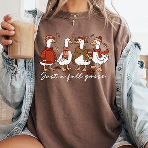 Peut inclure: T-shirt marron avec une illustration fantaisiste de quatre oies v&ecirc;tues de tenues d'automne. Les oies portent des pulls, des &eacute;charpes et des chapeaux, avec le texte "Just a fall goose" en &eacute;criture cursive. Le t-shirt est de style d&eacute;contract&eacute;.