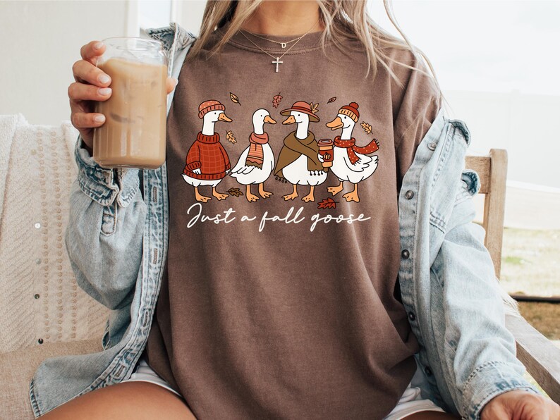 Peut inclure: T-shirt marron avec une illustration fantaisiste de quatre oies v&ecirc;tues de tenues d'automne. Les oies portent des pulls, des &eacute;charpes et des chapeaux, avec le texte "Just a fall goose" en &eacute;criture cursive. Le t-shirt est de style d&eacute;contract&eacute;.