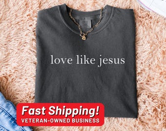 May include: A dark gray t-shirt with the text "love like jesus" printed in white. The shirt is laying on a fuzzy pink surface.