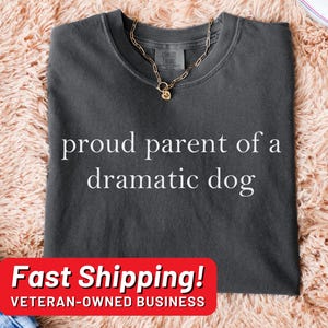May include: A dark gray t-shirt with the text "proud parent of a dramatic dog" printed in white. The shirt is laying on a light pink fuzzy surface. The text "Fast Shipping! VETERAN-OWNED BUSINESS" is printed in white on a red rectangle.