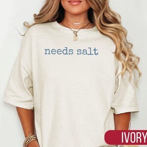 May include: Ivory-colored, oversized t-shirt with the text "needs salt" in blue. The shirt has a relaxed fit and short sleeves. The word "IVORY" is displayed in a red rectangle at the bottom of the image.