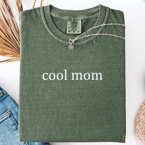 May include: A green t-shirt with the text "cool mom" printed in white on the front. The shirt is folded and laid flat on a white surface.