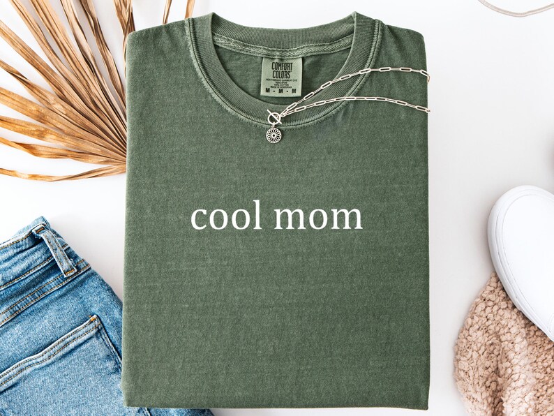 May include: A green t-shirt with the text "cool mom" printed in white on the front. The shirt is folded and laid flat on a white surface.