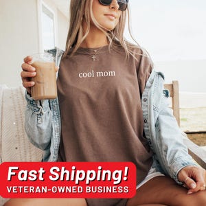 May include: A woman wearing a brown t-shirt with the text "cool mom" printed on it. She is sitting on a wooden chair and holding a glass of iced coffee. She is wearing a denim jacket and has sunglasses on.