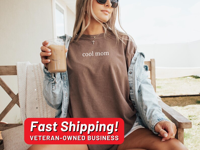 May include: A woman wearing a brown t-shirt with the text "cool mom" printed on it. She is sitting on a wooden chair and holding a glass of iced coffee. She is wearing a denim jacket and has sunglasses on.
