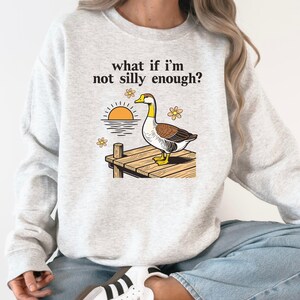 May include: Light gray sweatshirt with the text "what if I'm not silly enough?" above a graphic of a duck on a dock with a sunset and flowers. The duck has brown, white, yellow, and black details. The sweatshirt is a casual style.