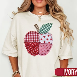 May include: An ivory short-sleeved t-shirt featuring a large, central apple graphic composed of four distinct patchwork-style fabric patterns in shades of red, white, and green, including gingham, floral, heart, and small apple prints.