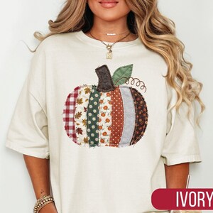May include: An ivory short-sleeved t-shirt featuring a large, central pumpkin graphic composed of vertical fabric strips in various patterns, including red gingham, autumn leaves, green floral, orange polka dots, and brown textures.