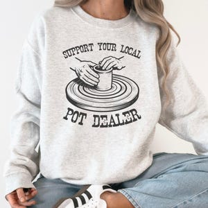 May include: Light gray sweatshirt with the text "SUPPORT YOUR LOCAL POT DEALER" in black. The design features a potter's wheel with hands shaping a pot. The sweatshirt is a casual, comfortable garment.