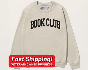 May include: A light beige sweatshirt with the text "BOOK CLUB" in black letters across the front. The sweatshirt has long sleeves and a crew neck.