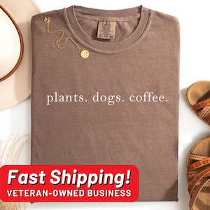 May include: A brown t-shirt with the text "plants. dogs. coffee." printed in white. The shirt has a crew neck and is made of a soft, comfortable fabric. The text "Fast Shipping! VETERAN-OWNED BUSINESS" is printed on a red banner at the bottom of the image.