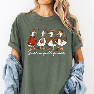 Peut inclure: T-shirt vert olive avec une illustration de quatre oies v&ecirc;tues de tenues d'automne. Chaque oie porte un chapeau, une &eacute;charpe ou un pull, et l'une d'elles tient une tasse de caf&eacute;. Le texte "Just a fall goose" est &eacute;crit en dessous.
