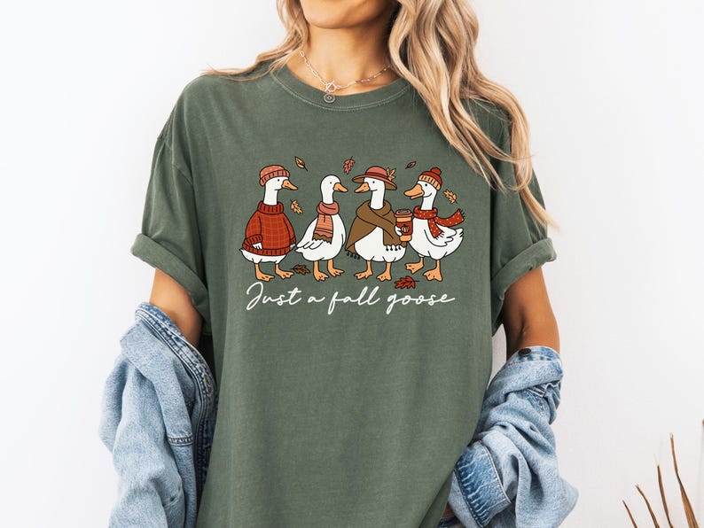 Peut inclure: T-shirt vert olive avec une illustration de quatre oies v&ecirc;tues de tenues d'automne. Chaque oie porte un chapeau, une &eacute;charpe ou un pull, et l'une d'elles tient une tasse de caf&eacute;. Le texte "Just a fall goose" est &eacute;crit en dessous.