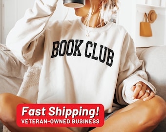 May include: Cream-colored sweatshirt with the text "BOOK CLUB" in bold, black, arched letters. The sweatshirt is long-sleeved and appears to be made of a soft material. The image shows a person wearing the sweatshirt.