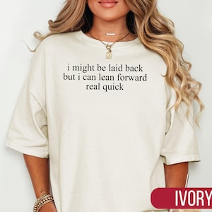 May include: Ivory-colored oversized t-shirt with the text "i might be laid back but i can lean forward real quick" in black. The shirt has a relaxed fit and short sleeves. The word "IVORY" is displayed in a red rectangle at the bottom.