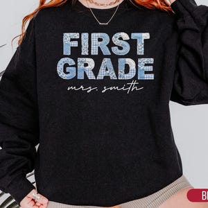 May include: A black long-sleeve sweatshirt featuring the text FIRST GRADE in a light blue and white patchwork pattern. Below the main text, the name mrs. smith is printed in a white cursive script font. The garment has a crew neckline.