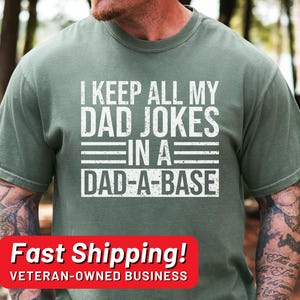 May include: Sage green t-shirt with the text "I KEEP ALL MY DAD JOKES IN A DAD-A-BASE" in white block letters. The shirt is a short-sleeved crew neck style.