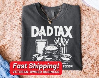 May include: A dark gray t-shirt with a white graphic of a burger, fries, and a soda. The text "DAD TAX" is printed in white above the graphic. The shirt has a distressed look.