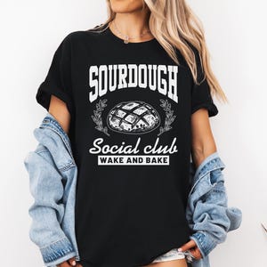 May include: Black t-shirt with white text that reads "Sourdough Social Club Wake and Bake". A drawing of a sourdough bread loaf is above the text.