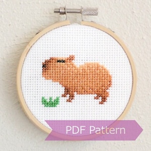 May include: A cross-stitch pattern of a brown capybara eating a green leaf, framed in a wooden embroidery hoop. The text "PDF Pattern" is printed on a purple ribbon below the hoop.